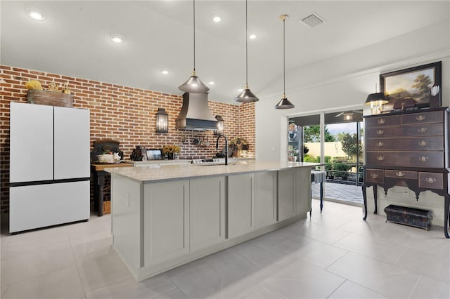 kitchen with light stone countertops, white refrigerator, premium range hood, an island with sink, and decorative light fixtures