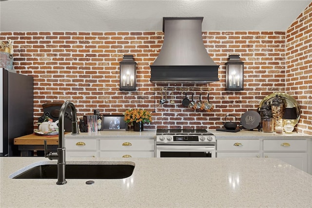 kitchen with appliances with stainless steel finishes, brick wall, sink, wall chimney range hood, and white cabinets