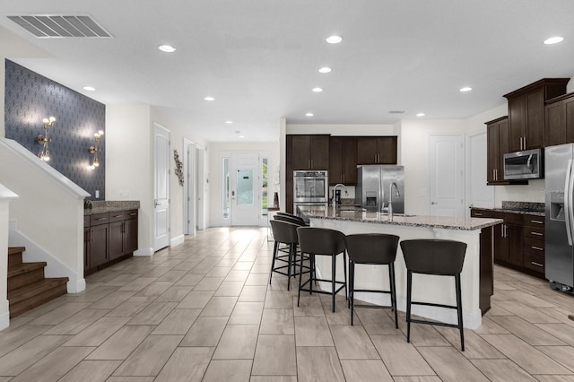 kitchen with stone counters, a center island with sink, dark brown cabinets, a kitchen bar, and stainless steel appliances