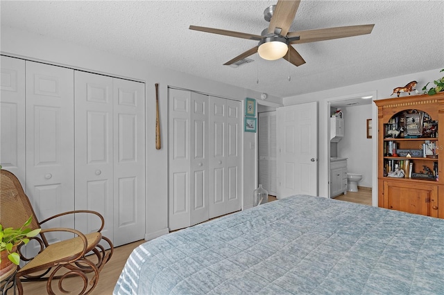 bedroom featuring two closets, light hardwood / wood-style flooring, ensuite bath, a textured ceiling, and ceiling fan