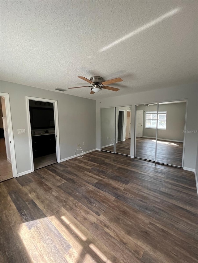 unfurnished bedroom with a textured ceiling, two closets, ceiling fan, and dark wood-type flooring