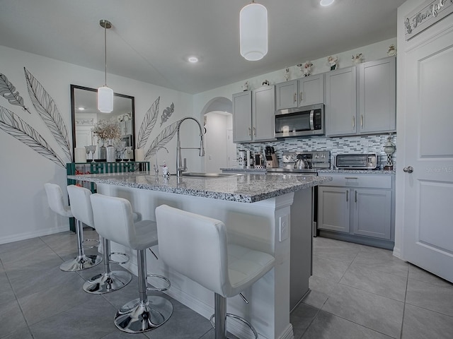 kitchen with gray cabinetry, stainless steel appliances, hanging light fixtures, and an island with sink