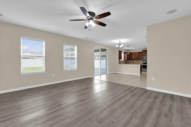 unfurnished living room with plenty of natural light, baseboards, light wood-style flooring, and ceiling fan with notable chandelier