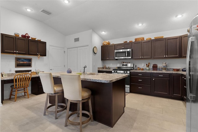 kitchen with a breakfast bar, a kitchen island with sink, vaulted ceiling, appliances with stainless steel finishes, and dark brown cabinets