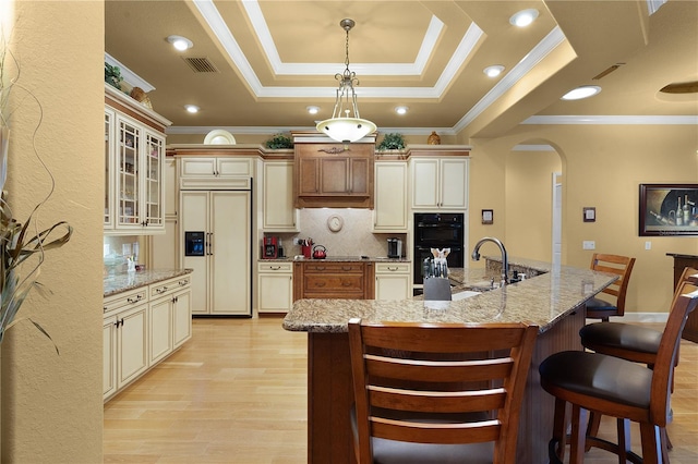 kitchen featuring a raised ceiling, tasteful backsplash, a large island with sink, cream cabinets, and paneled built in refrigerator