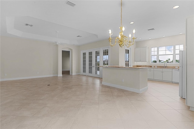 kitchen with ceiling fan with notable chandelier, a tray ceiling, a kitchen island, light tile patterned floors, and white cabinetry