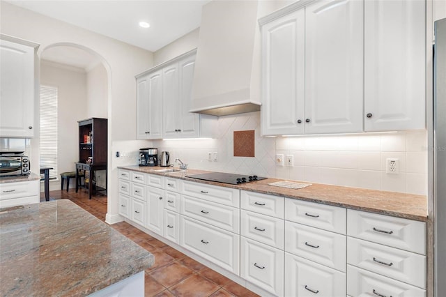 kitchen with black electric stovetop, backsplash, premium range hood, stone counters, and white cabinets