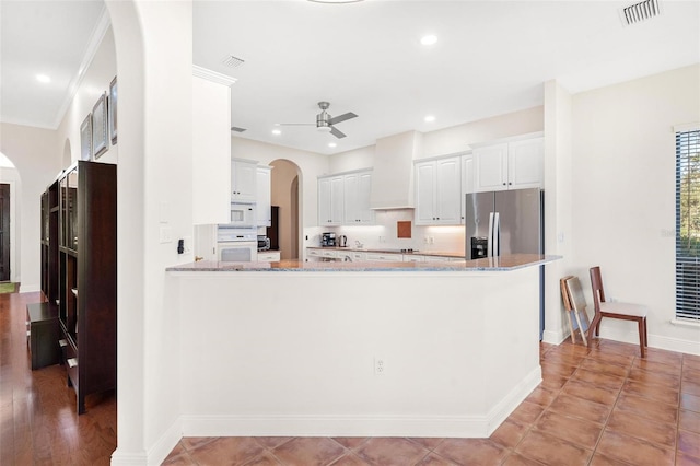 kitchen featuring wall chimney range hood, white appliances, light stone counters, white cabinets, and kitchen peninsula