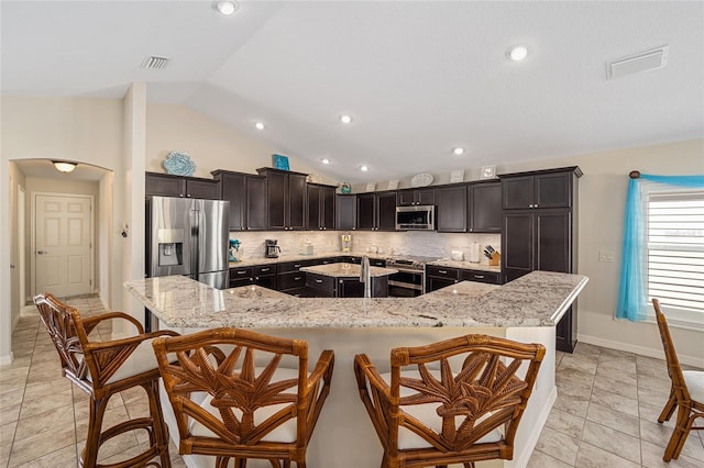 kitchen with light stone countertops, tasteful backsplash, a large island with sink, vaulted ceiling, and appliances with stainless steel finishes