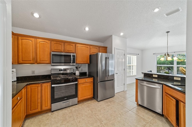 kitchen with light tile patterned floors, appliances with stainless steel finishes, hanging light fixtures, a textured ceiling, and dark stone counters