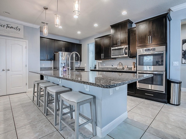 kitchen with dark stone counters, a center island with sink, hanging light fixtures, ornamental molding, and appliances with stainless steel finishes