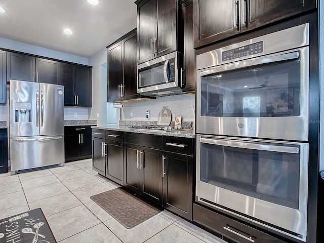 kitchen featuring light stone countertops, stainless steel appliances, tasteful backsplash, dark brown cabinets, and light tile patterned floors