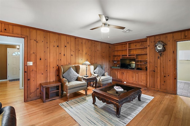 living room featuring built in shelves, ceiling fan, light hardwood / wood-style floors, and wood walls