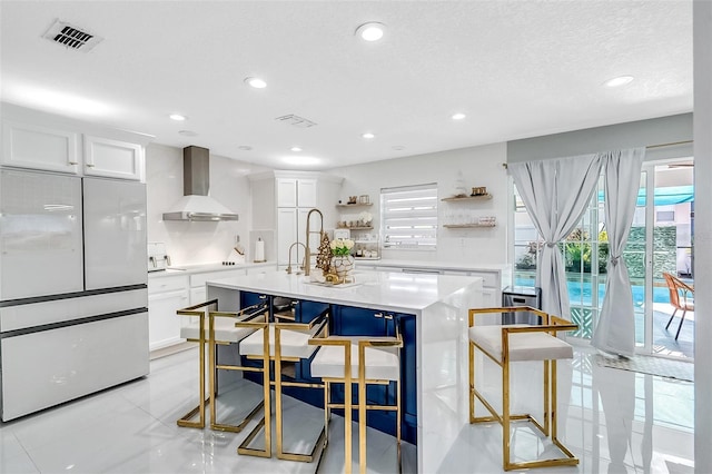 kitchen featuring white cabinetry, wall chimney range hood, built in refrigerator, an island with sink, and a breakfast bar