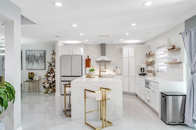 kitchen featuring a kitchen bar, wall chimney exhaust hood, sink, white cabinetry, and an island with sink