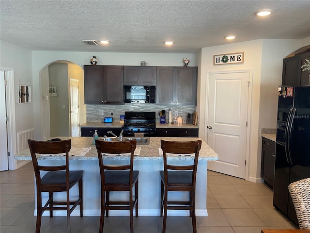 kitchen featuring black appliances, a center island with sink, light stone countertops, light tile patterned floors, and a textured ceiling