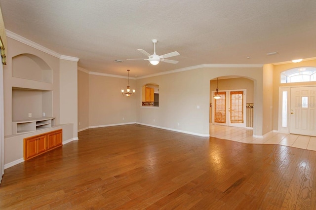 unfurnished living room with ceiling fan with notable chandelier, built in features, crown molding, and light hardwood / wood-style flooring