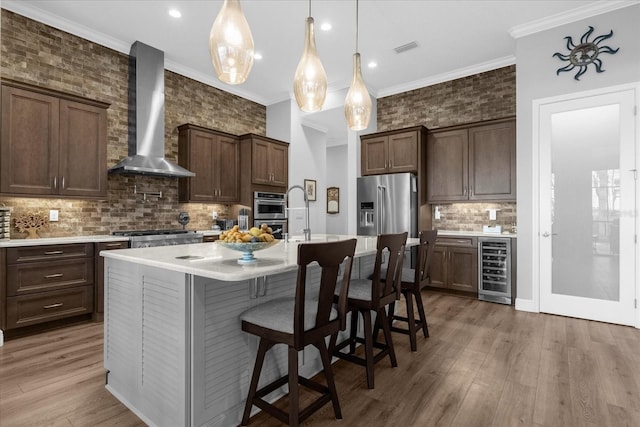 kitchen featuring stainless steel appliances, beverage cooler, wall chimney range hood, pendant lighting, and an island with sink