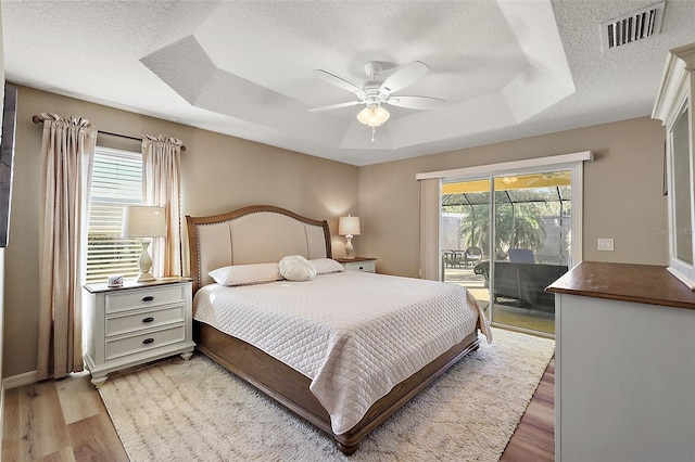 bedroom with a textured ceiling, access to outside, a raised ceiling, ceiling fan, and light hardwood / wood-style flooring