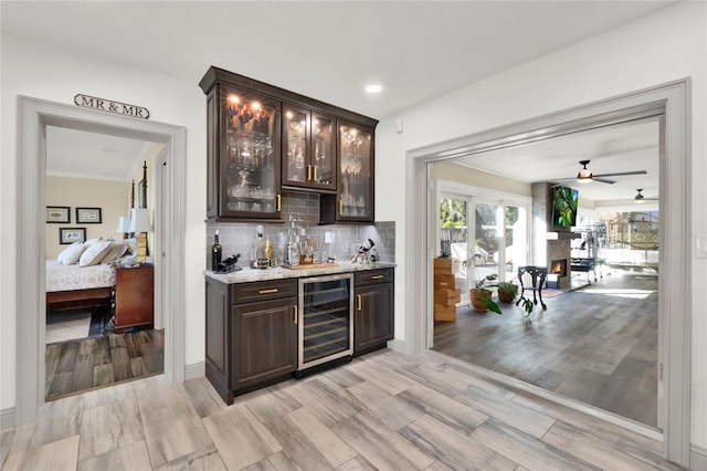 bar with light wood-type flooring, backsplash, dark brown cabinets, ceiling fan, and wine cooler