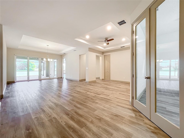 unfurnished living room with french doors, ceiling fan with notable chandelier, a tray ceiling, and light hardwood / wood-style floors