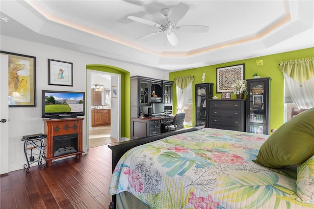 bedroom with a raised ceiling, connected bathroom, ceiling fan, and dark wood-type flooring