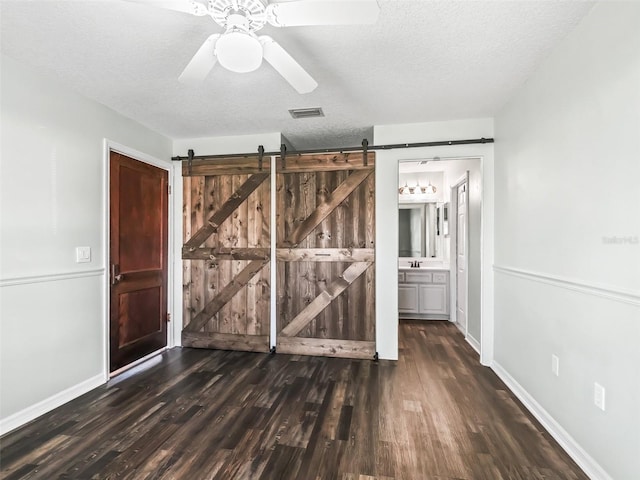 unfurnished bedroom with ensuite bath, ceiling fan, a barn door, a textured ceiling, and dark hardwood / wood-style flooring