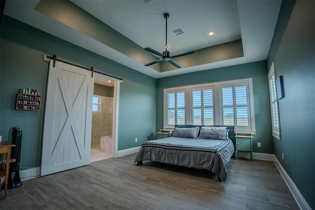 bedroom with a tray ceiling, ceiling fan, a barn door, hardwood / wood-style floors, and connected bathroom