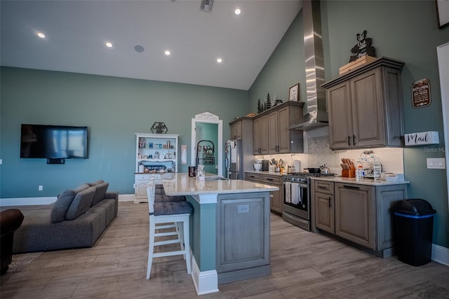 kitchen featuring wall chimney range hood, a kitchen breakfast bar, high vaulted ceiling, an island with sink, and appliances with stainless steel finishes