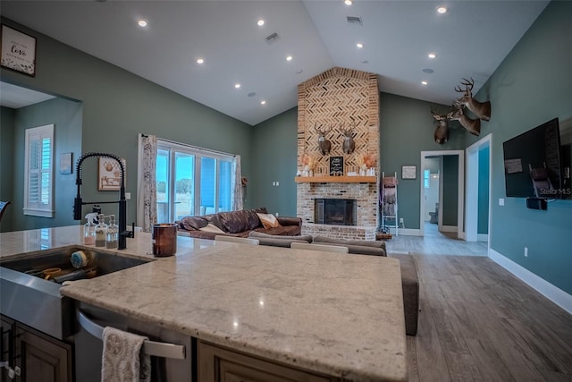 kitchen with light wood-type flooring, a brick fireplace, light stone counters, sink, and high vaulted ceiling