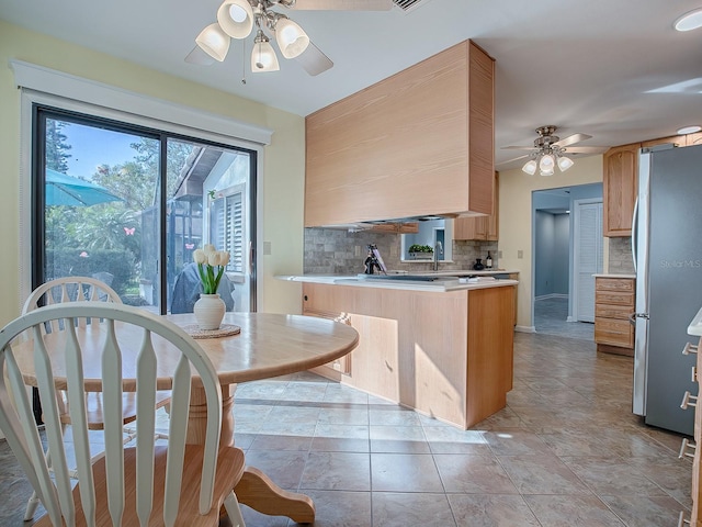 kitchen with kitchen peninsula, backsplash, ceiling fan, light brown cabinets, and stainless steel refrigerator