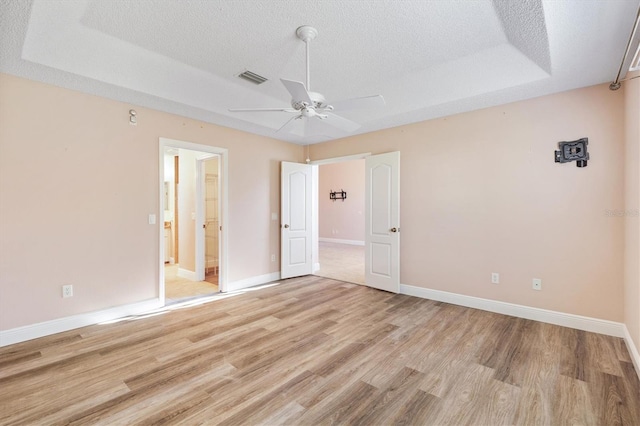 unfurnished bedroom with ensuite bath, ceiling fan, light hardwood / wood-style floors, a textured ceiling, and a tray ceiling