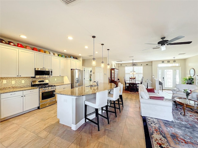 kitchen with appliances with stainless steel finishes, light stone counters, decorative light fixtures, white cabinets, and an island with sink