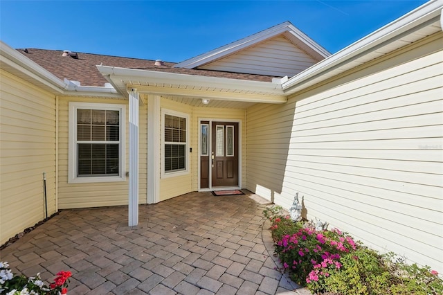 view of exterior entry with roof with shingles and a patio