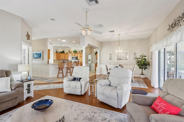 living room featuring ceiling fan with notable chandelier, wood finished floors, visible vents, and ornate columns