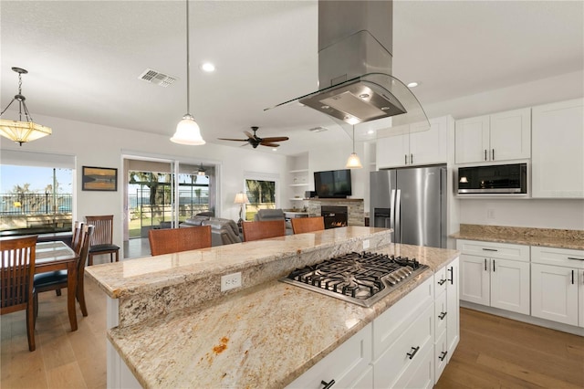 kitchen featuring appliances with stainless steel finishes, island range hood, a kitchen island, a stone fireplace, and hanging light fixtures