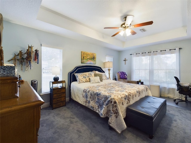 bedroom with multiple windows, dark carpet, a raised ceiling, and ceiling fan