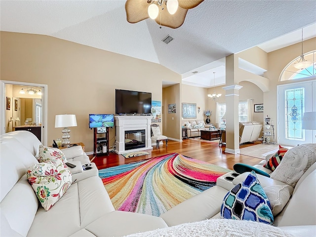 living room with ceiling fan with notable chandelier, vaulted ceiling, light wood-type flooring, a textured ceiling, and decorative columns