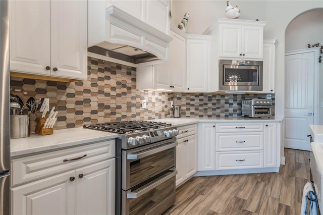 kitchen with custom exhaust hood, light wood-type flooring, appliances with stainless steel finishes, tasteful backsplash, and white cabinetry