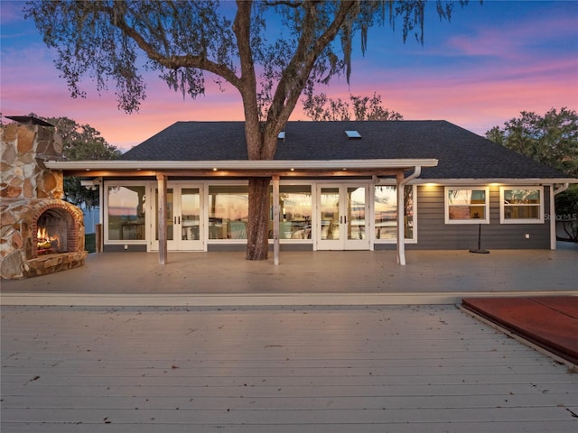 back house at dusk with a deck, french doors, and an outdoor stone fireplace