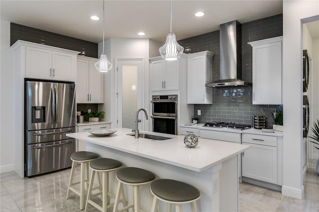 kitchen with pendant lighting, a center island with sink, wall chimney range hood, appliances with stainless steel finishes, and white cabinetry