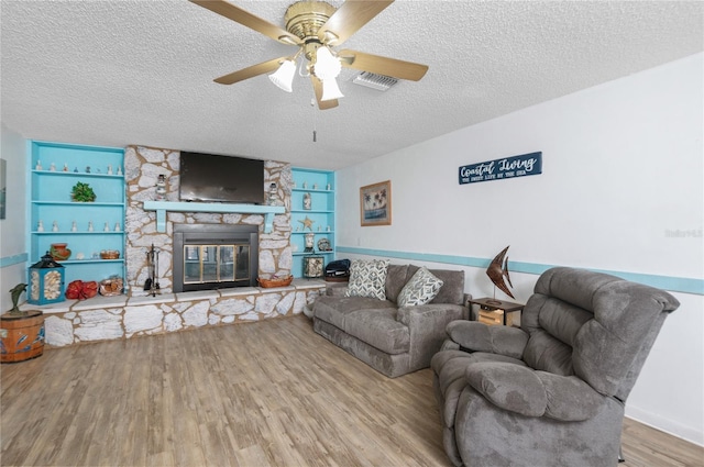 living room featuring hardwood / wood-style floors, a stone fireplace, built in shelves, ceiling fan, and a textured ceiling