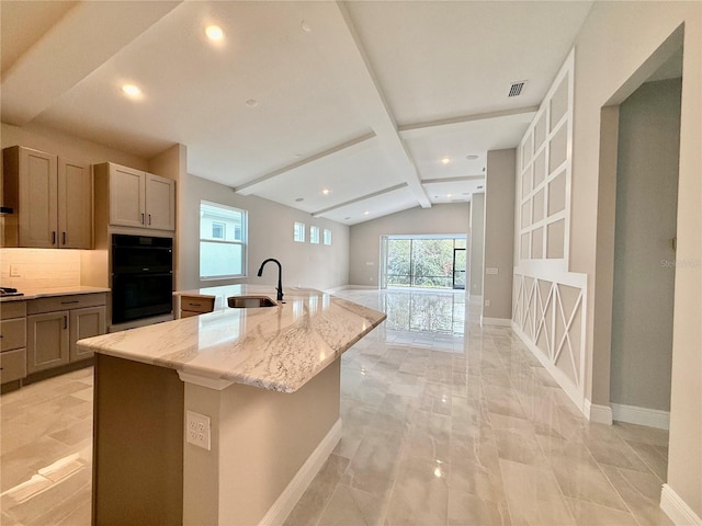 kitchen with light stone countertops, black double oven, sink, lofted ceiling with beams, and a center island with sink