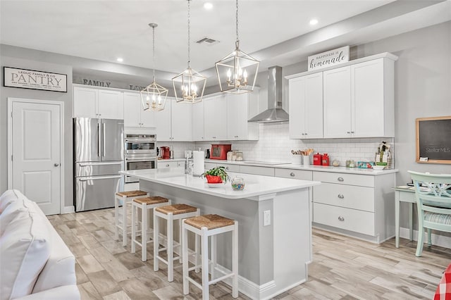 kitchen with decorative light fixtures, white cabinetry, wall chimney range hood, stainless steel appliances, and a center island with sink