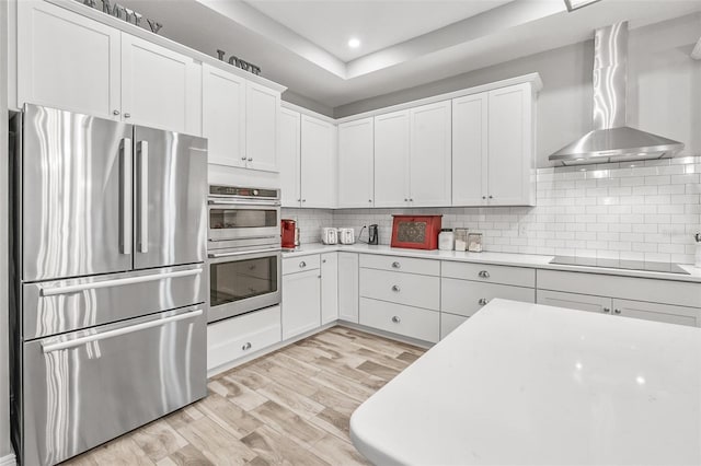 kitchen featuring backsplash, a tray ceiling, stainless steel appliances, white cabinets, and wall chimney exhaust hood