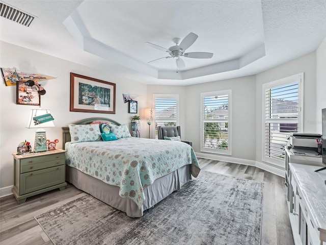 bedroom with a textured ceiling, ceiling fan, light hardwood / wood-style floors, and a tray ceiling