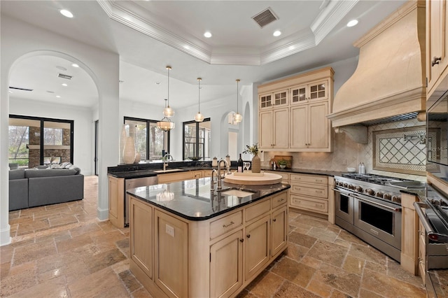 kitchen with appliances with stainless steel finishes, an island with sink, decorative light fixtures, a tray ceiling, and custom exhaust hood