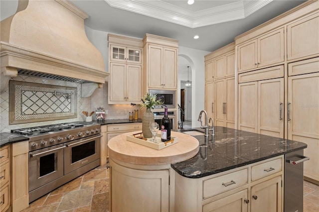 kitchen featuring an island with sink, stainless steel appliances, ornamental molding, custom range hood, and a raised ceiling