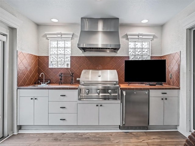 kitchen featuring tile countertops, sink, white cabinets, stainless steel fridge, and wall chimney exhaust hood