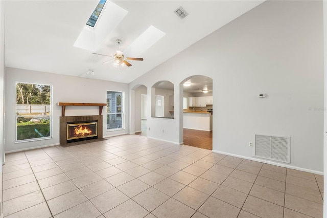 unfurnished living room featuring light tile patterned floors, ceiling fan, vaulted ceiling with skylight, and a brick fireplace